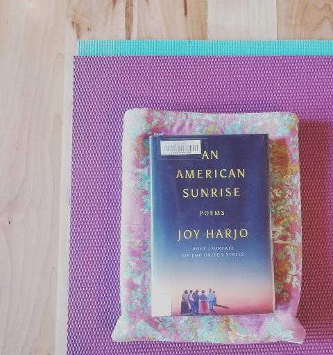 Image of An American Sunrise: Poems by Joy Harjo on a pink, yellow, turquoise book sleeve on a purple and turquoise yoga mat on a wood floor