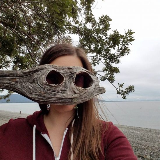 Melissa With Long Brown Hair On A Beach Holding A Piece Of Drift Wood With Two Holes That Looks Like A Wooden Mask.