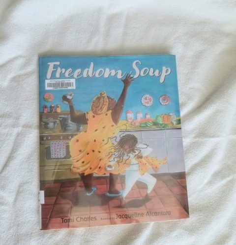 The Illustrated Kids Book Freedom Soup On A Linen Cloth Background. The Book Shows A Haitian Grandmother In A Yellow Dress And A Little Haitian Girl In White Dancing With A Spice Shaker In A Kitchen.