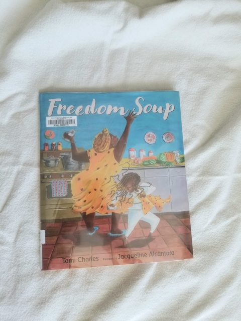 The illustrated kids book Freedom Soup on a linen cloth background. The book shows a Haitian grandmother in a yellow dress and a little Haitian girl in white dancing with a spice shaker in a kitchen.